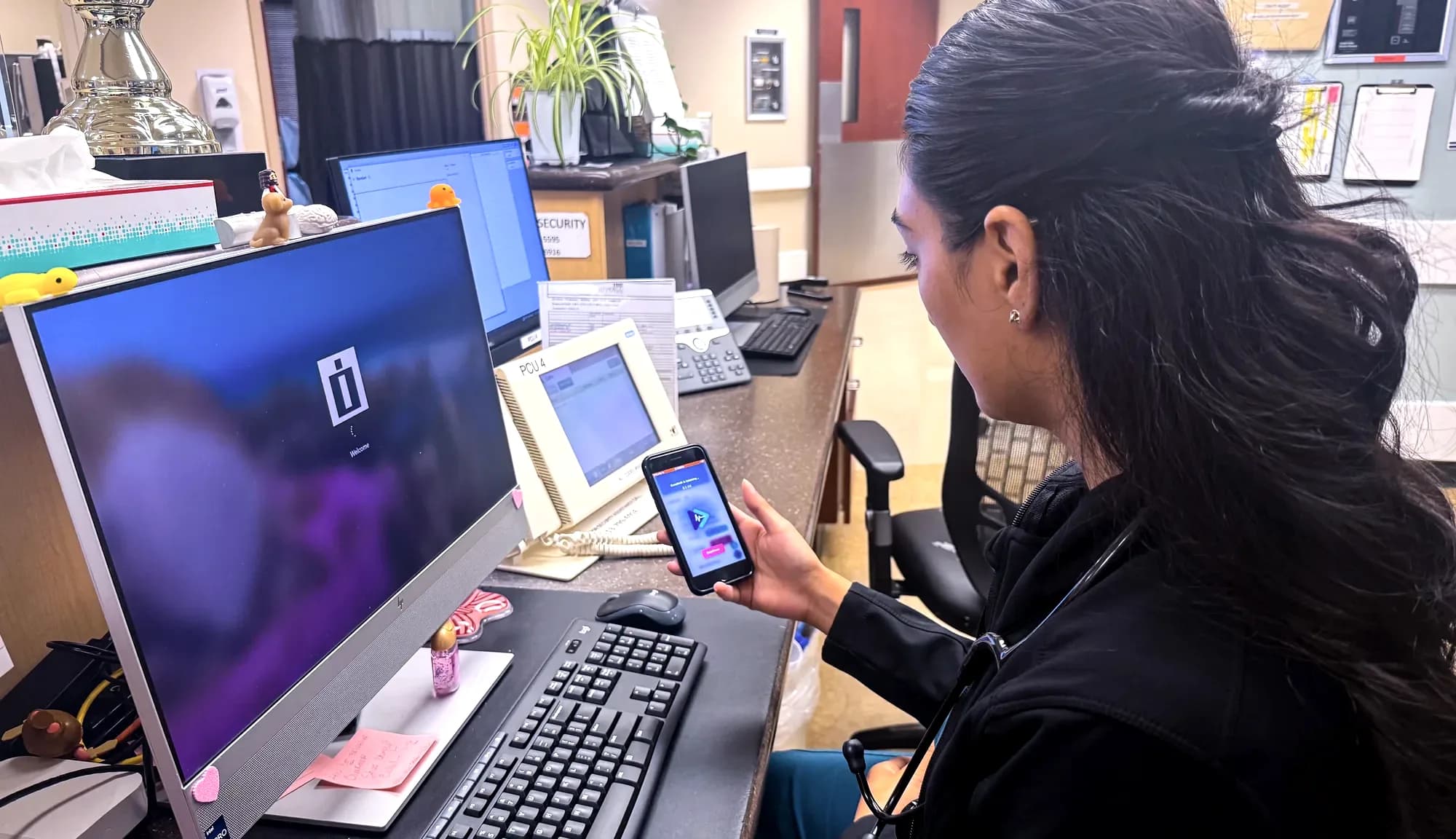 A nurse at the station holding a phone running CareShift beside her computer